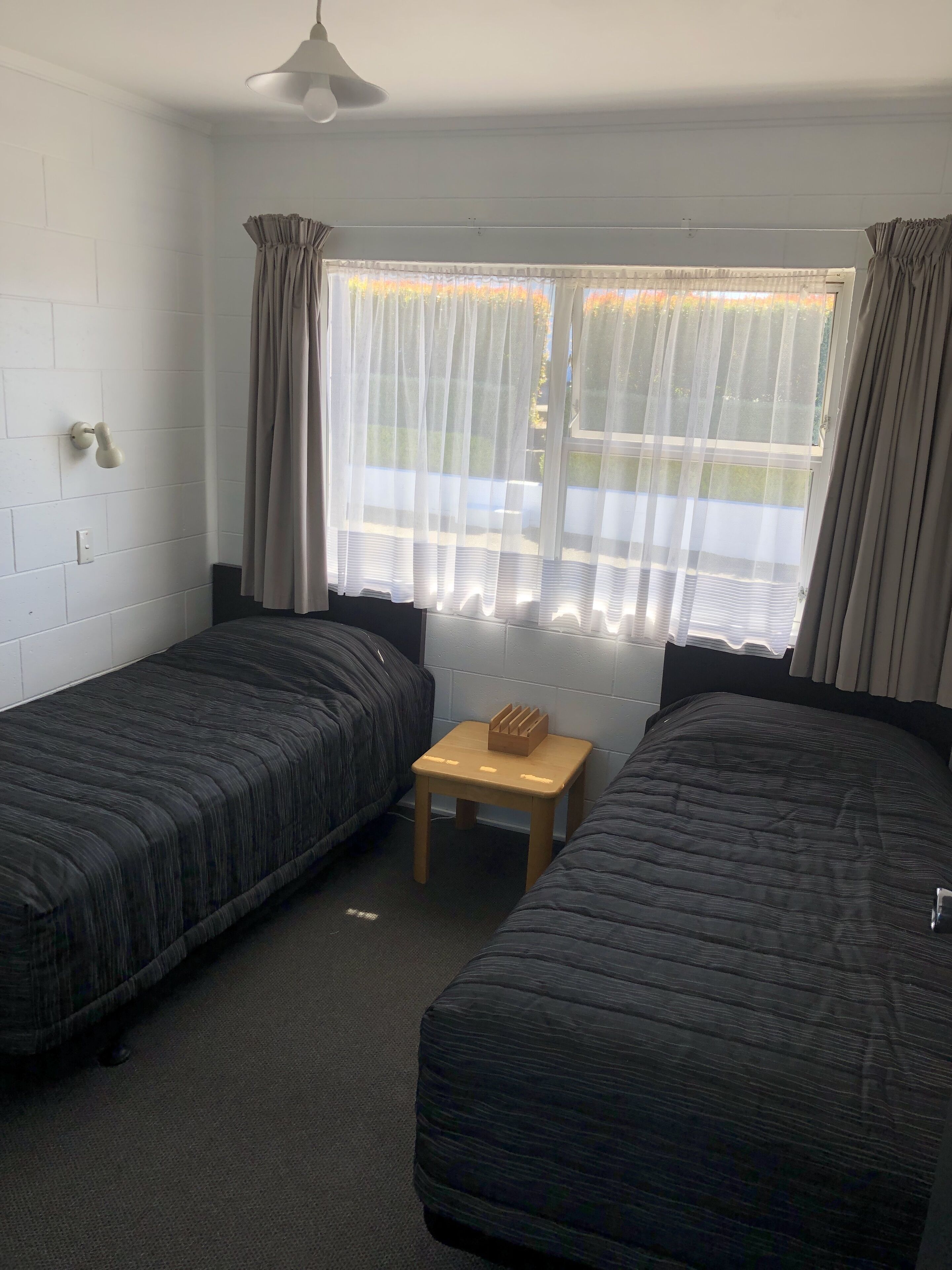 Quarto com duas camas de solteiro, uma pequena mesa de madeira, ventoinha no teto e janela com cortinas finas.