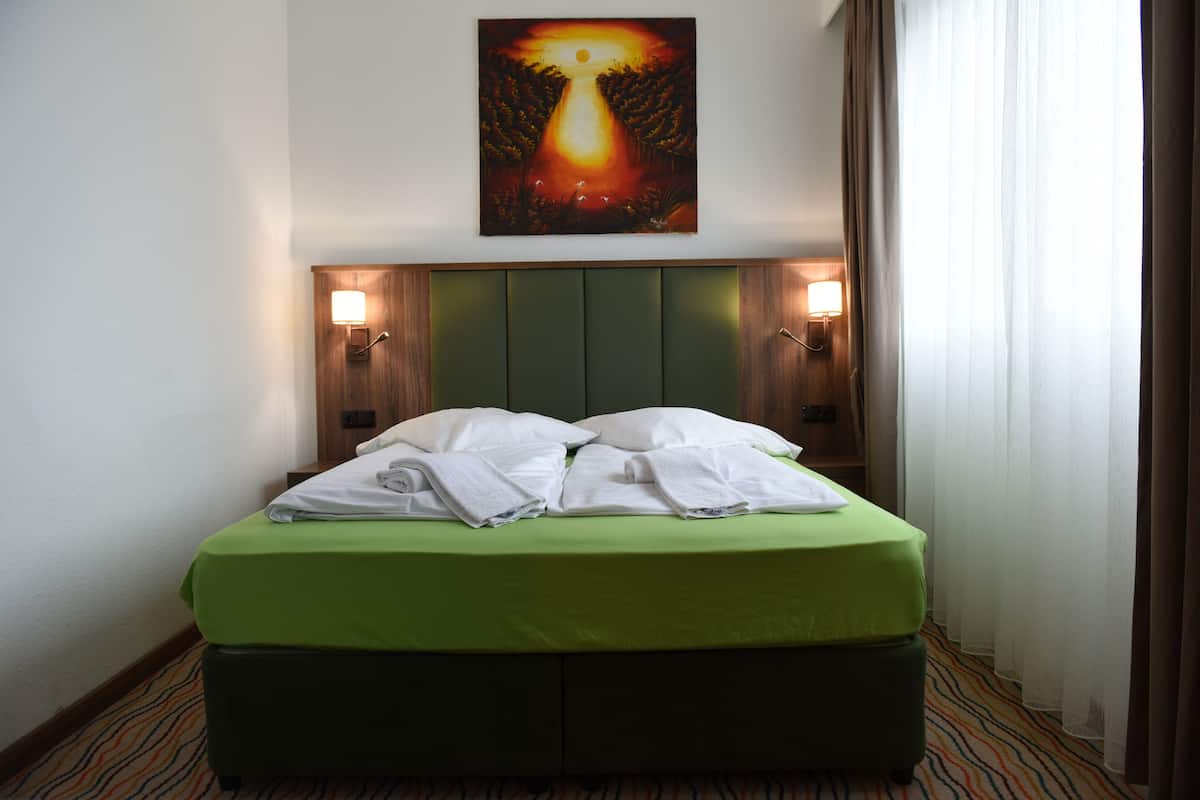 Habitación de hotel con una cama verde, dos lámparas de noche, un cuadro sobre la cama y una ventana con cortinas.
