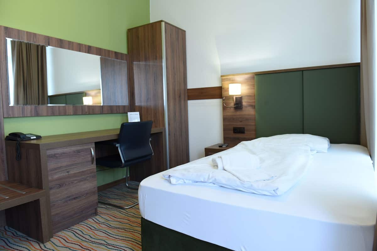 Habitación de hotel con escritorio de madera, una silla, cama con ropa blanca, espejo y mesita de noche con lámpara.