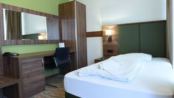 Habitación de hotel con escritorio de madera, una silla, cama con ropa blanca, espejo y mesita de noche con lámpara.