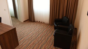 Una habitación de hotel con alfombra estampada, un escritorio de madera, un sillón negro y vistas al exterior a través de una ventana con cortinas.