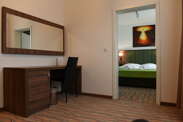 A hotel room with a wooden desk, a chair, a bed with green bedding, a patterned carpet, a large mirror, and a painting on the wall.