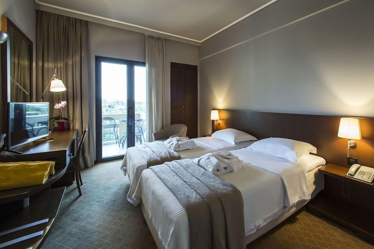 A hotel room with two beds, a desk with a computer, a chair, and a balcony with a view.