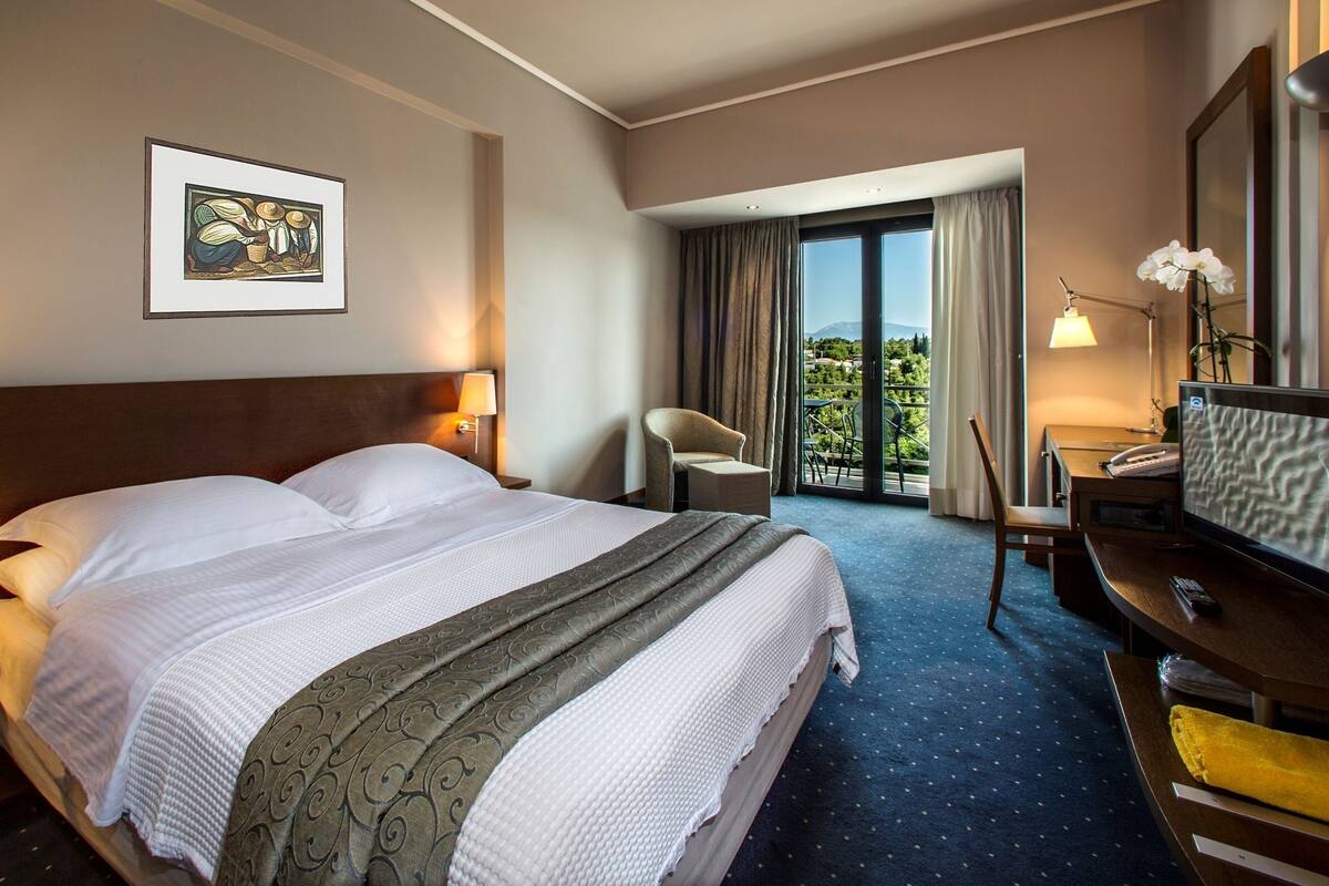 A hotel room with a large bed, a desk with a TV, a chair, and a balcony with curtains.