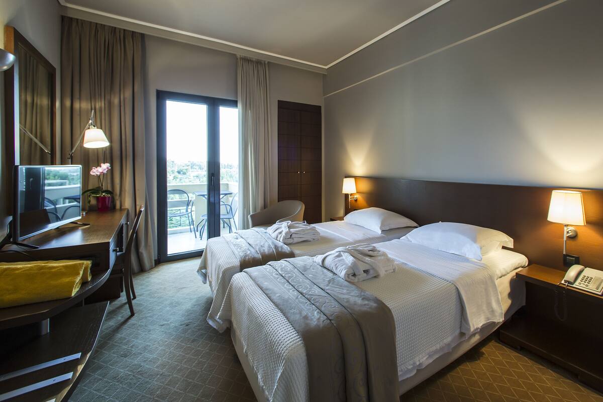 A hotel room with two beds, a desk with a computer, a chair, and a view of the outdoors through a sliding door.