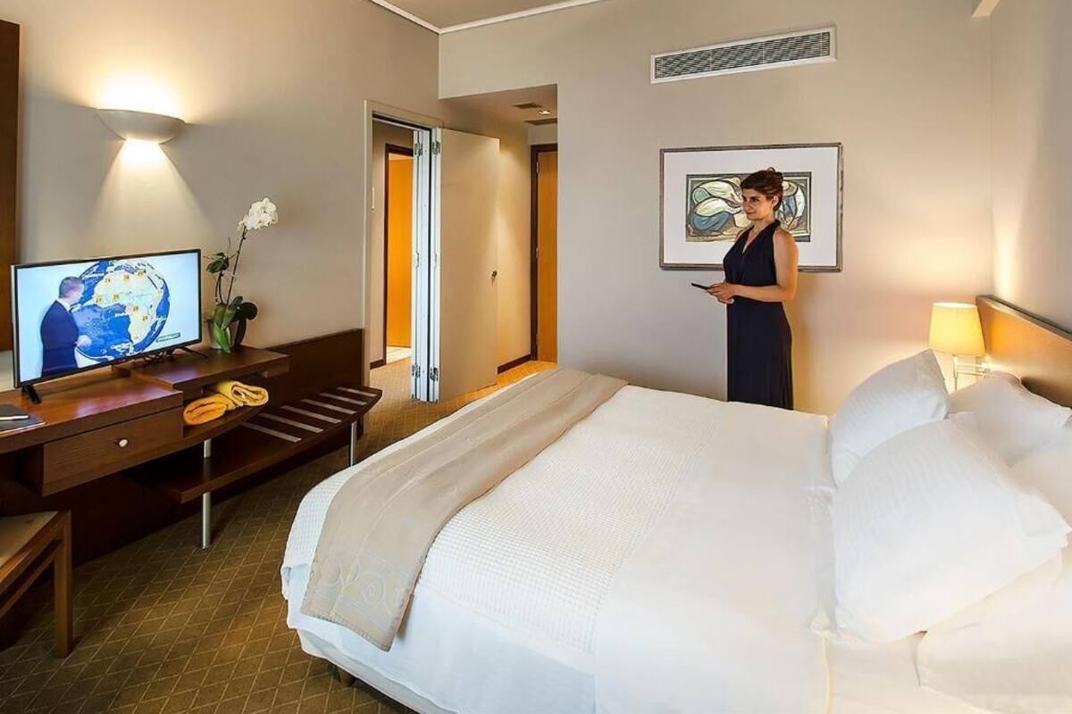 A hotel room with a bed, a desk with a television, a standing lamp, and a door leading to another room.