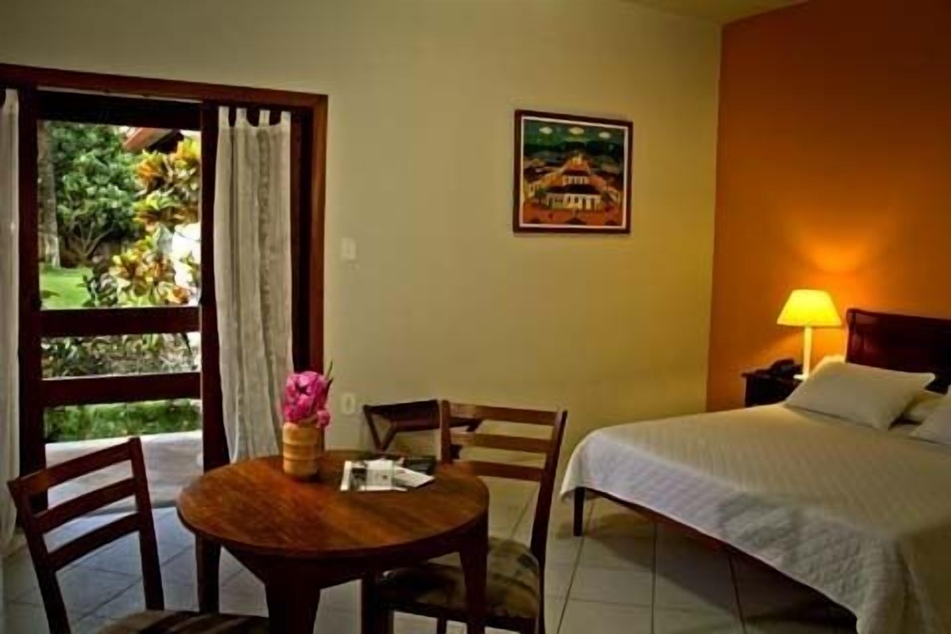 Quarto de hotel com cama, mesa com cadeiras, janela com cortinas, um quadro na parede e abajur na mesa de cabeceira.