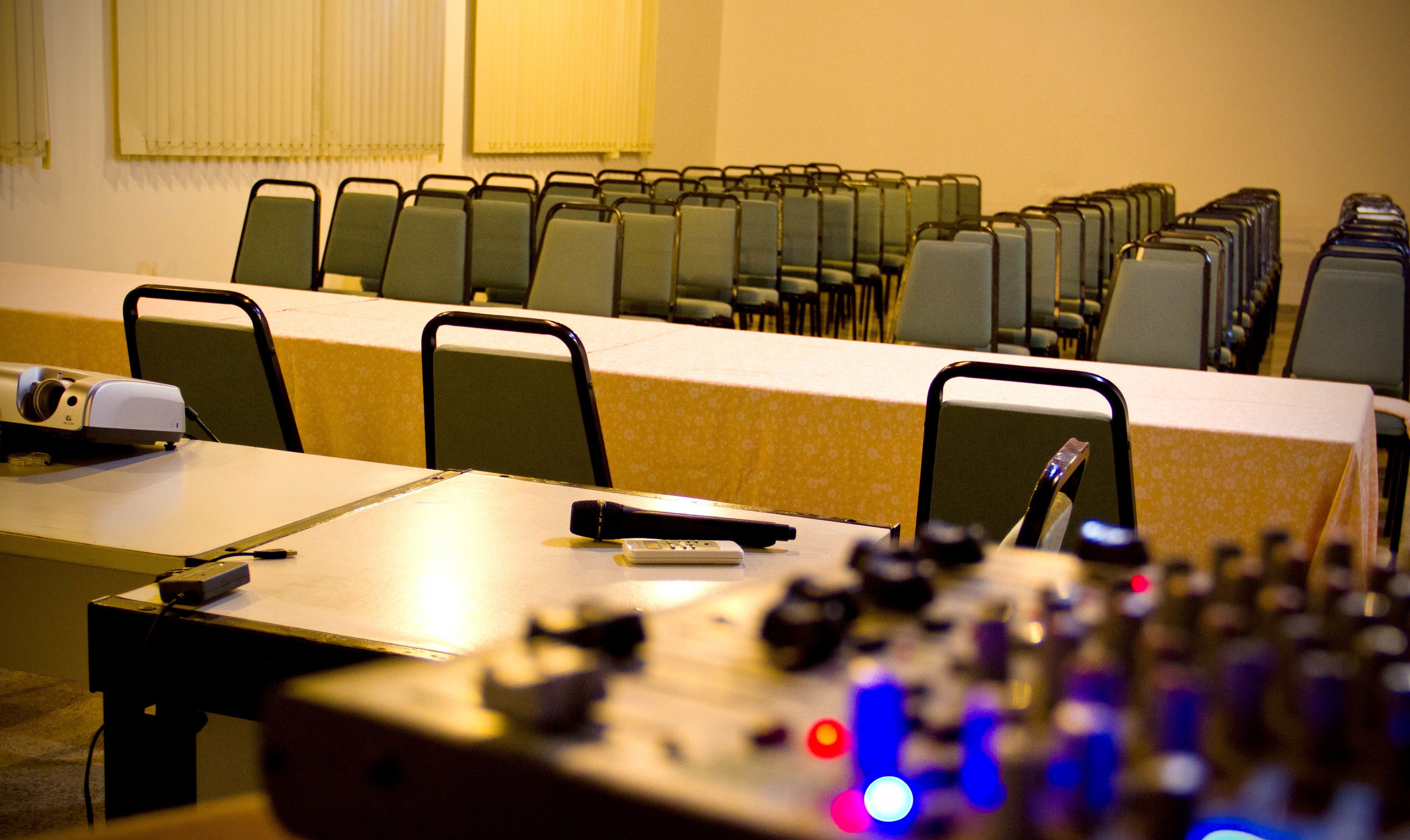 Un salón de conferencias con filas de sillas, una mesa con proyector y un micrófono.