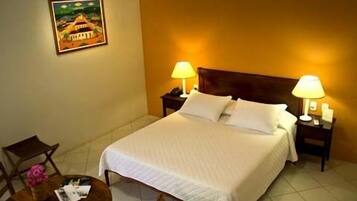 Una habitación de hotel con una cama, dos lámparas de noche, una mesita de noche, una pequeña mesa con un jarrón y un cuadro en la pared.
