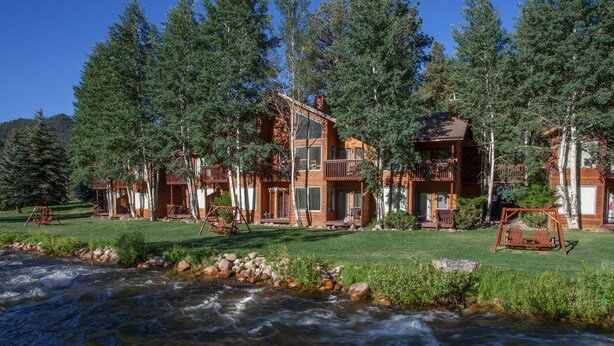 A lodge with multiple wooden cabins, a river in the foreground, and a forested backdrop. - Aspen Winds on Fall River (Estes Park)