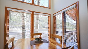 A dining area with a wooden table and chairs, large windows with wooden blinds, and a pine cone decoration on the table. - Aspen Winds on Fall River (Estes Park)