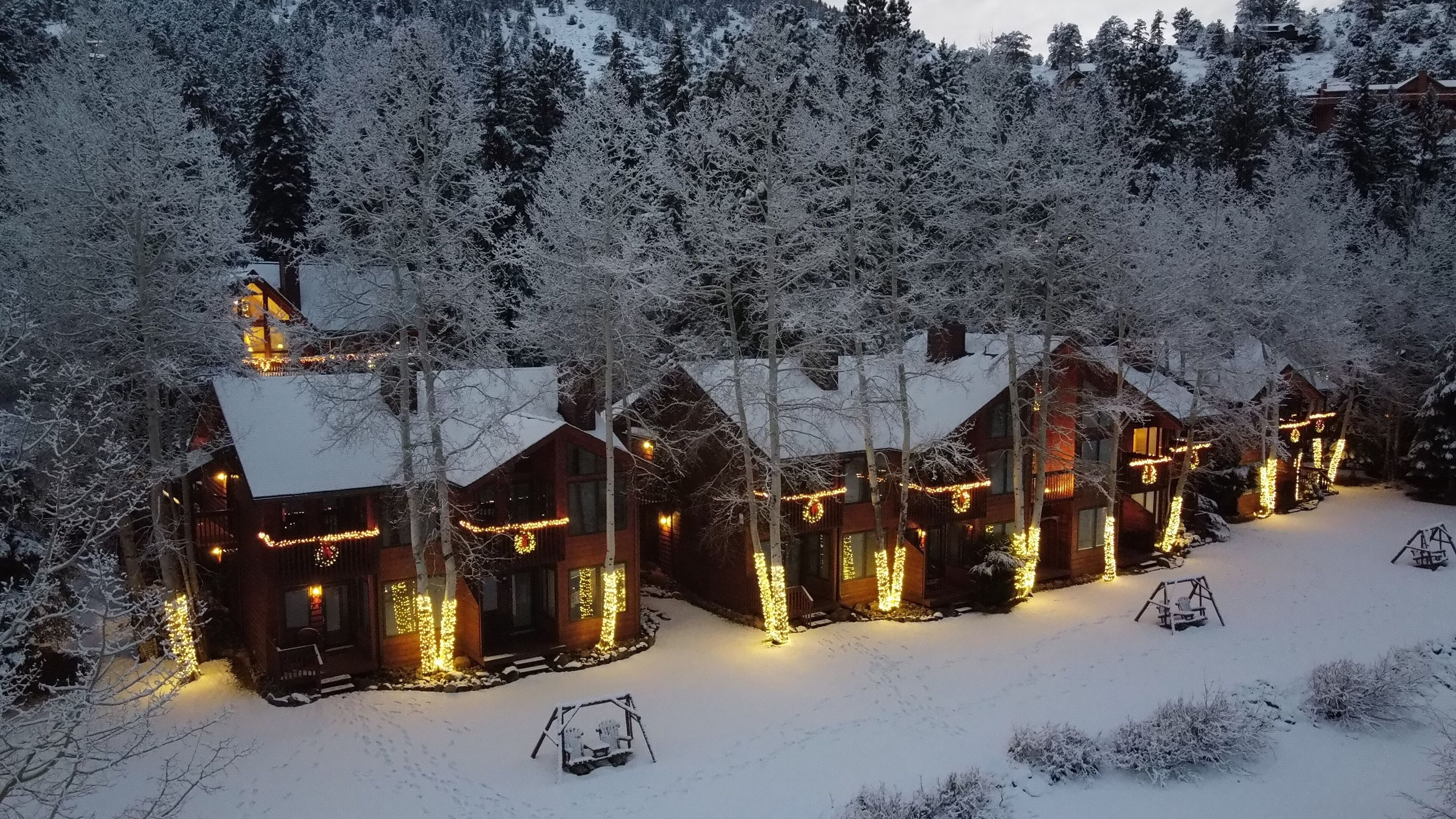 Filas de cabañas de madera con tejados nevados y luces de cuerda iluminadas, rodeadas por un bosque cubierto de nieve.
