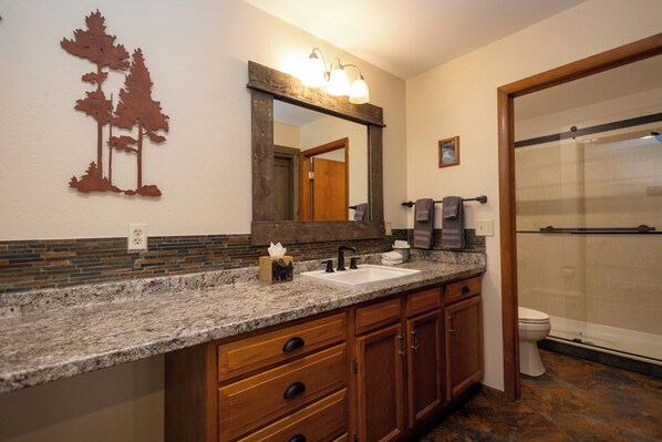 A bathroom with a granite countertop, wooden cabinets, a large mirror, and a glass shower enclosure.