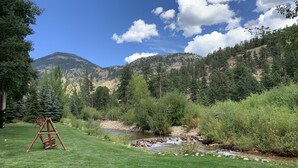 A grassy area with a swing set, a small stream, and a forested mountain backdrop. - Aspen Winds on Fall River (Estes Park)