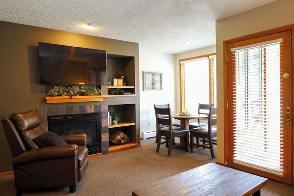 A living room with a fireplace, a leather chair, a wooden coffee table, and a dining area with a table and chairs. - Aspen Winds on Fall River (Estes Park)