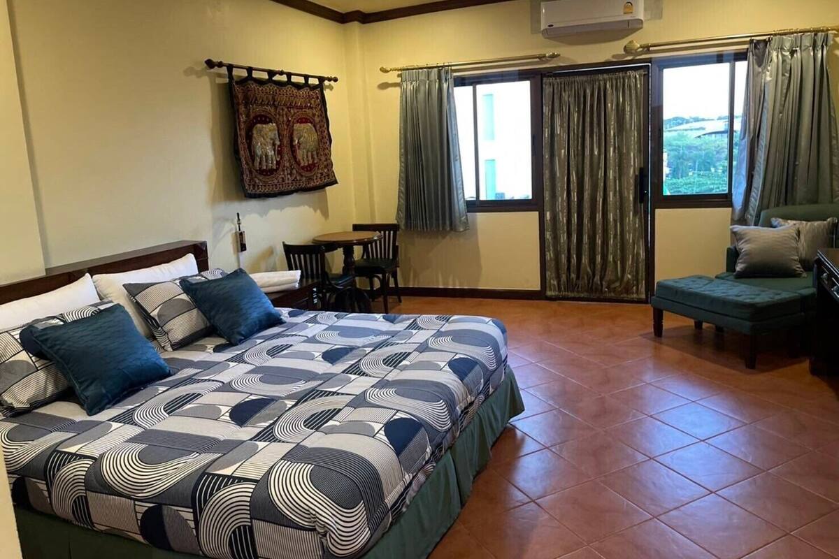 Un dormitorio con cama, escritorio, silla y sofá. Se aprecia una ventana con cortinas, un equipo de aire acondicionado y un cuadro colgado en la pared.