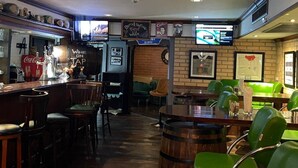 Un espace bar avec un comptoir, des tabourets et un écran de télévision. Des tableaux encadrés ornent le mur, et une enseigne indique : « Vin en fût, vin rouge, vin blanc ».