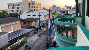 Une vue urbaine mettant en valeur un bâtiment imposant, doté d’un balcon verdoyant et d’un mât sur lequel flottent plusieurs drapeaux.