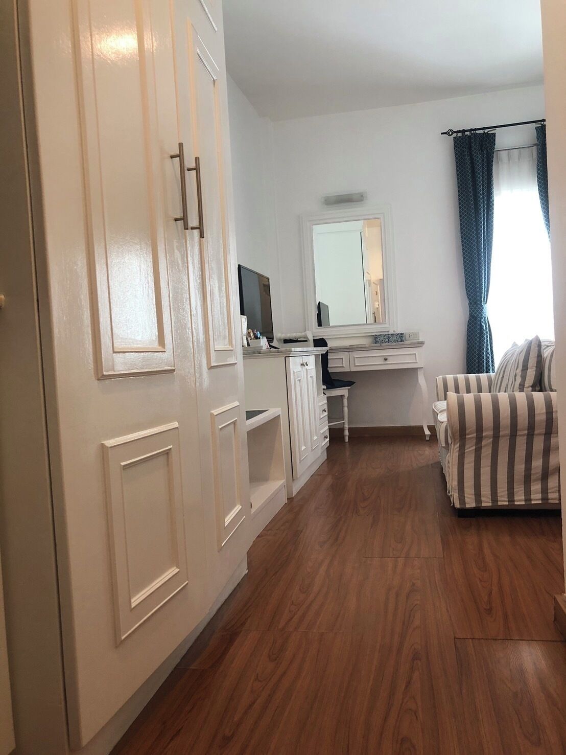 A bedroom with a wooden floor, a bed, a dresser, a desk with a mirror, and a window with curtains.