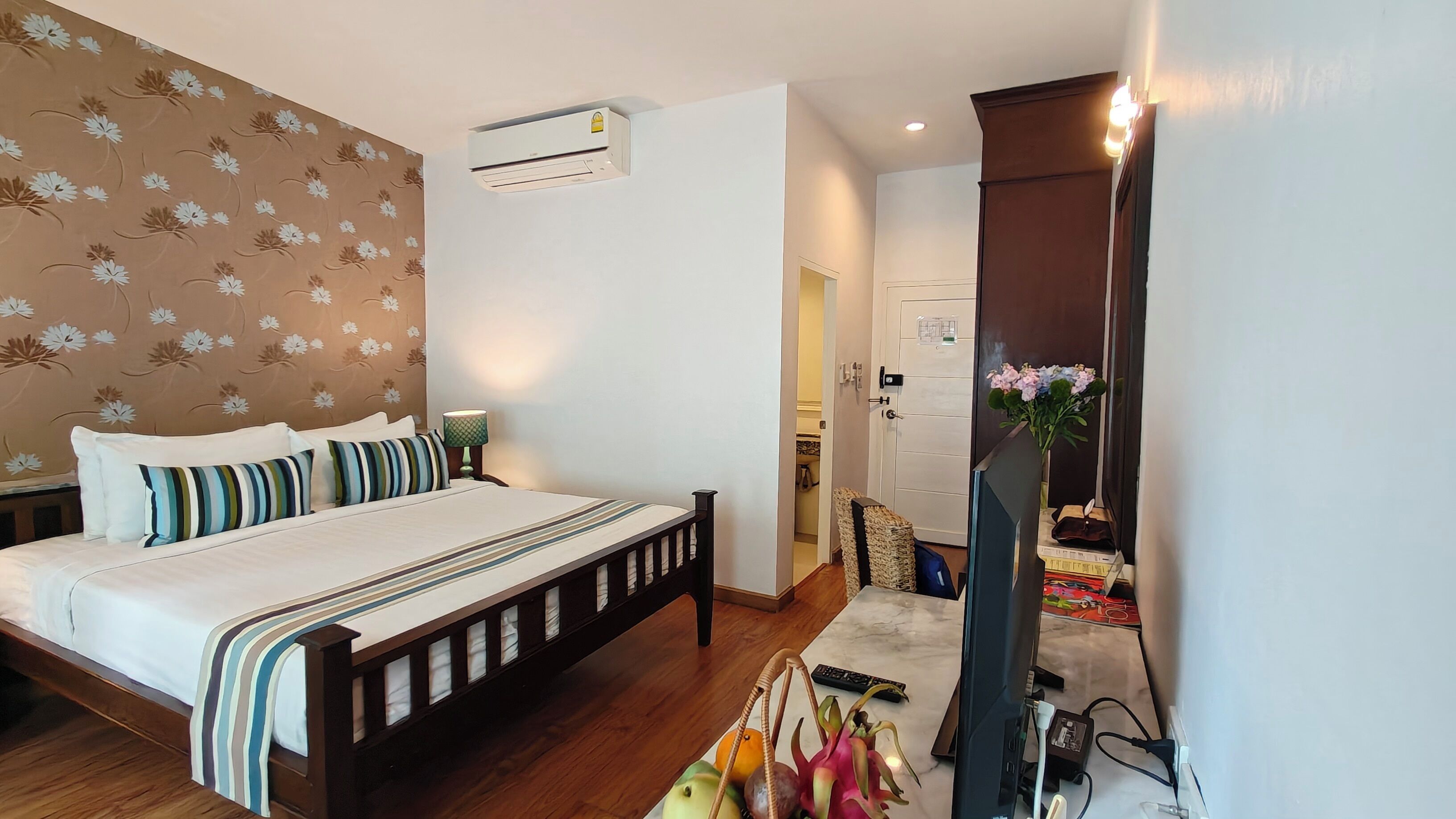 A bedroom with a bed, bedside lamp, air conditioning unit, and a TV stand with a vase of flowers.