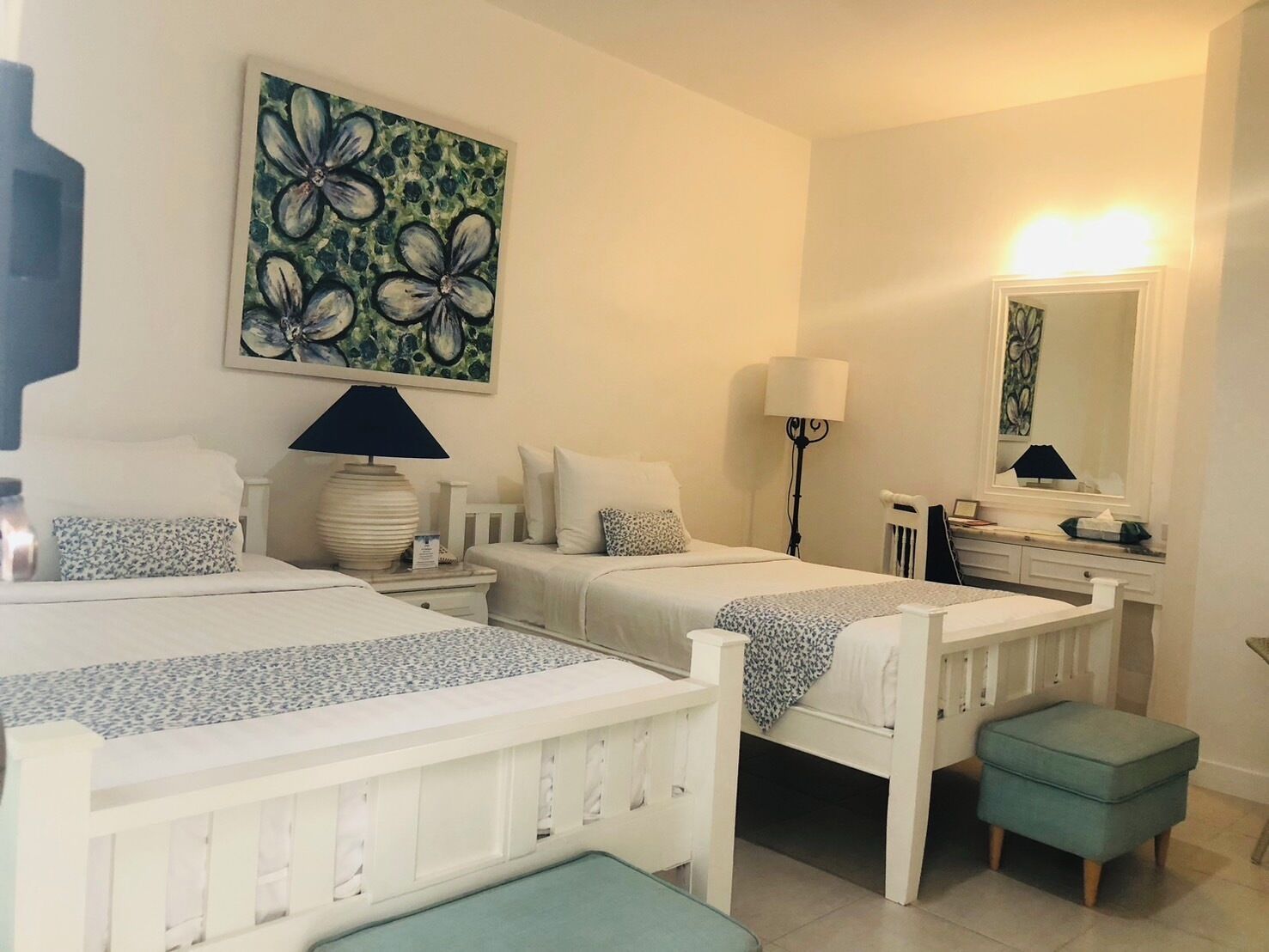 A hotel room with two single beds, a small desk, a mirror, a floor lamp, and a painting on the wall.