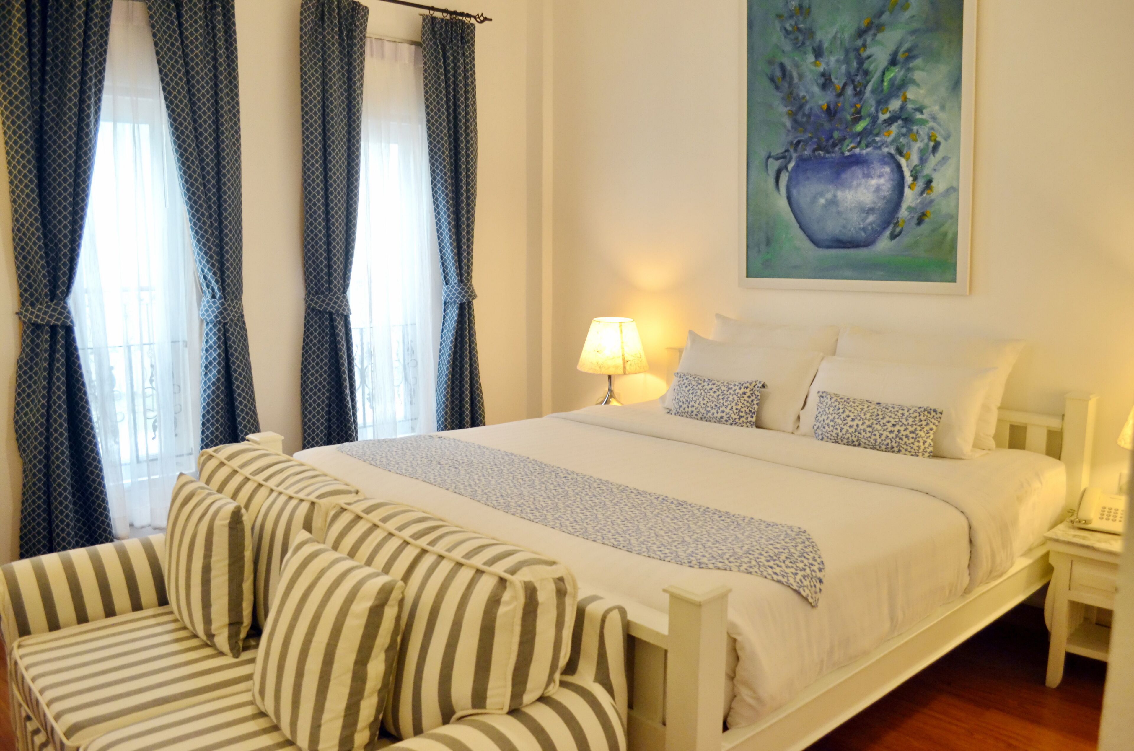 A hotel room with a bed, a bedside table with a lamp, a striped sofa, and a painting on the wall.