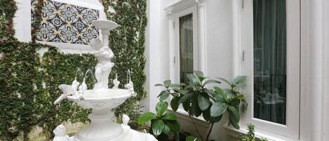 A white fountain with decorative figures, surrounded by lush greenery and a patterned tile wall.