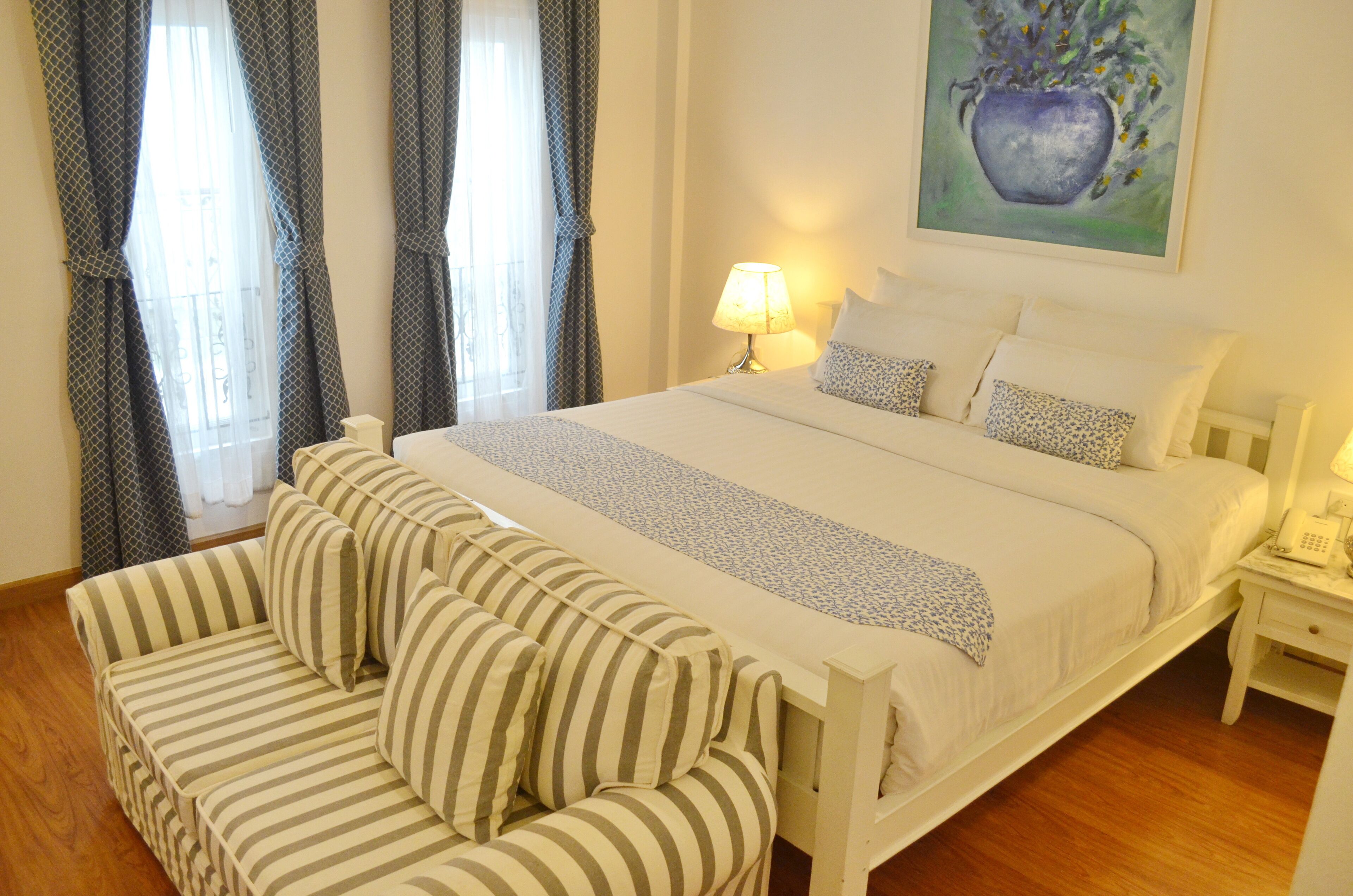 A hotel room with a bed, a sofa, a lamp, a painting, and a telephone on a nightstand.