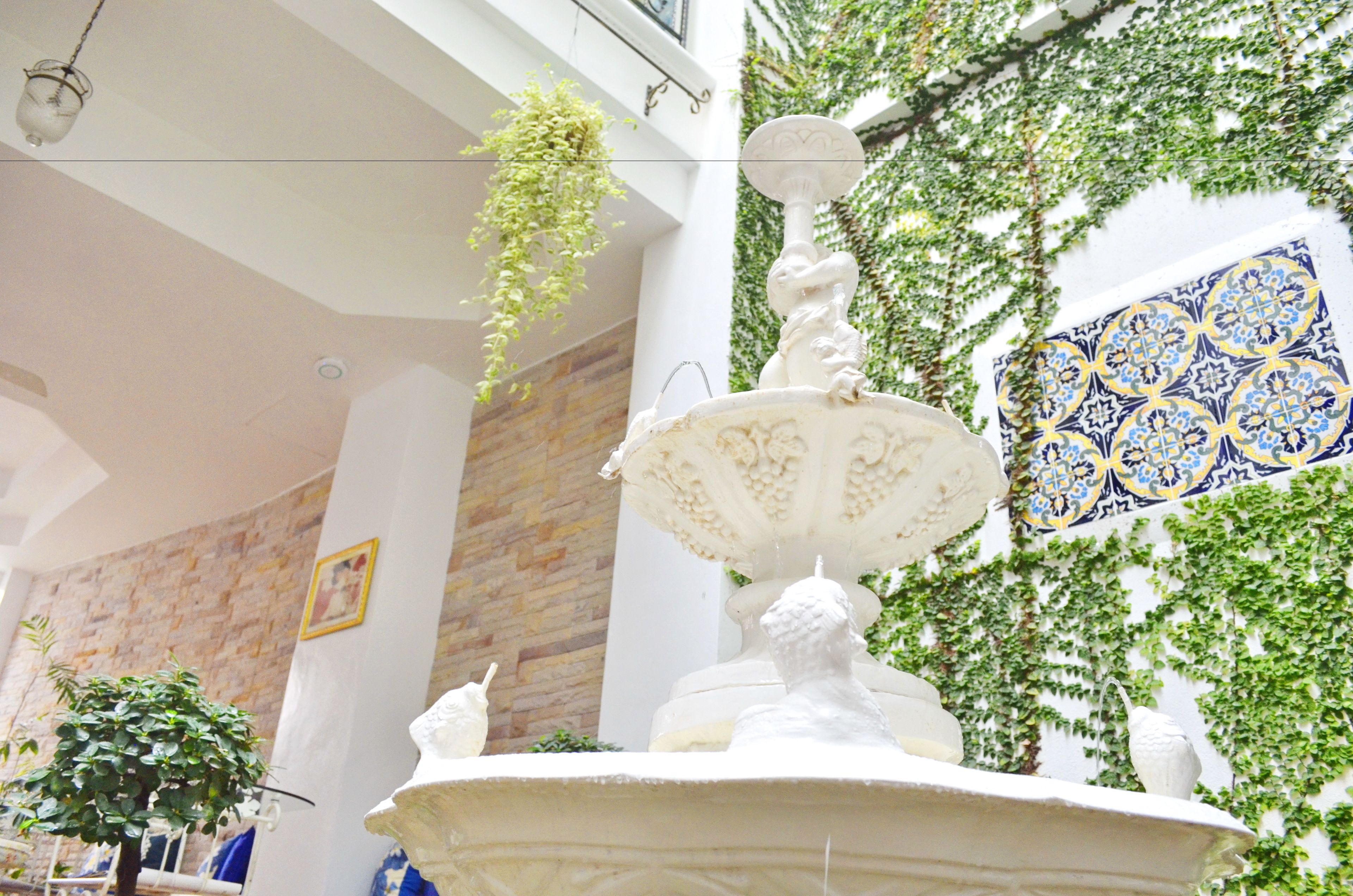 A fountain with a statue in a room with a brick wall, a hanging plant, and a patterned wall.