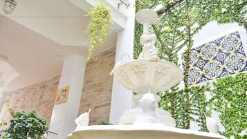 A fountain with a statue in a room with a brick wall, a hanging plant, and a patterned wall.
