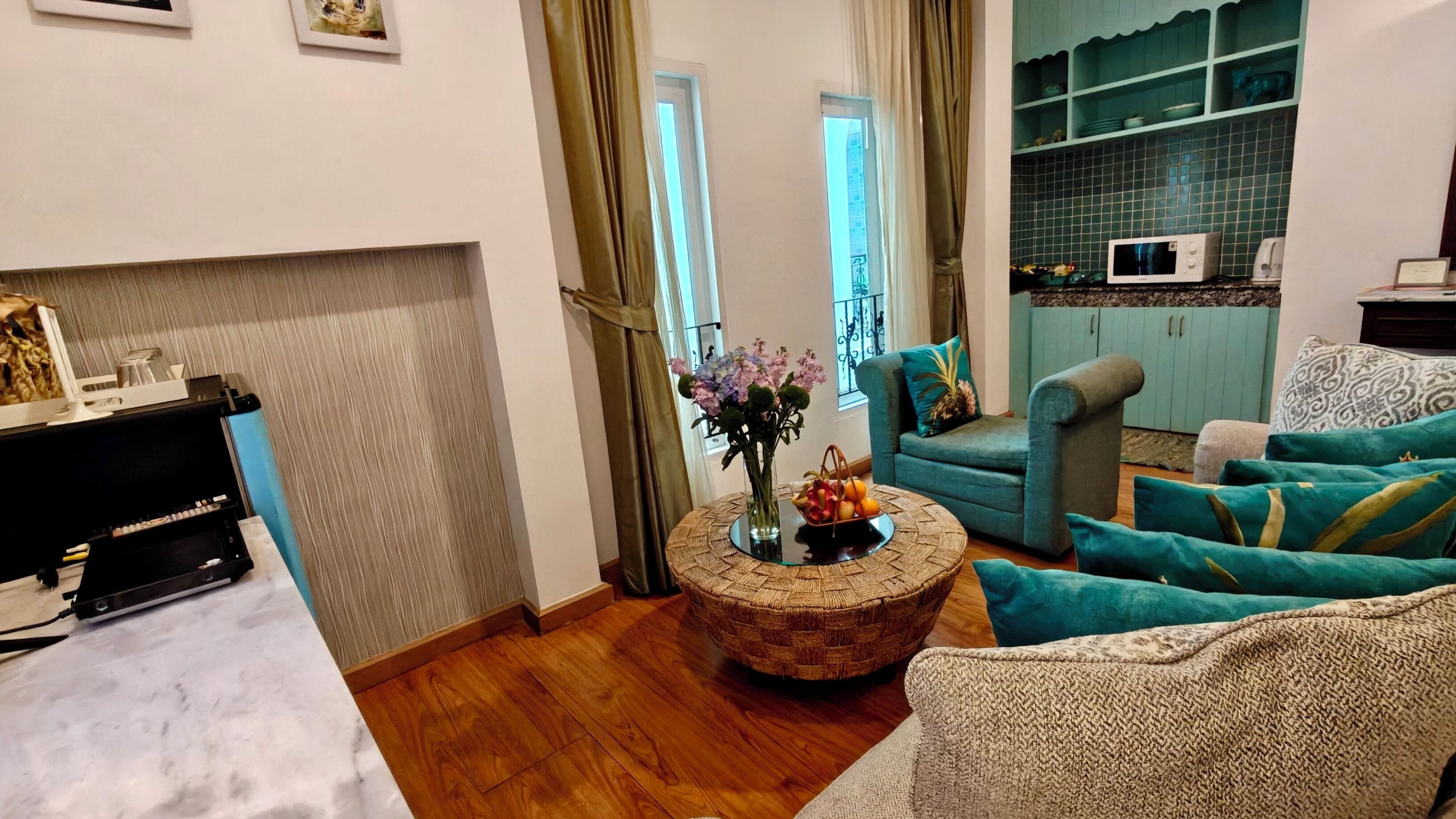 A living room with a sofa set, a coffee table with flowers, a TV, and a kitchen area with a microwave.