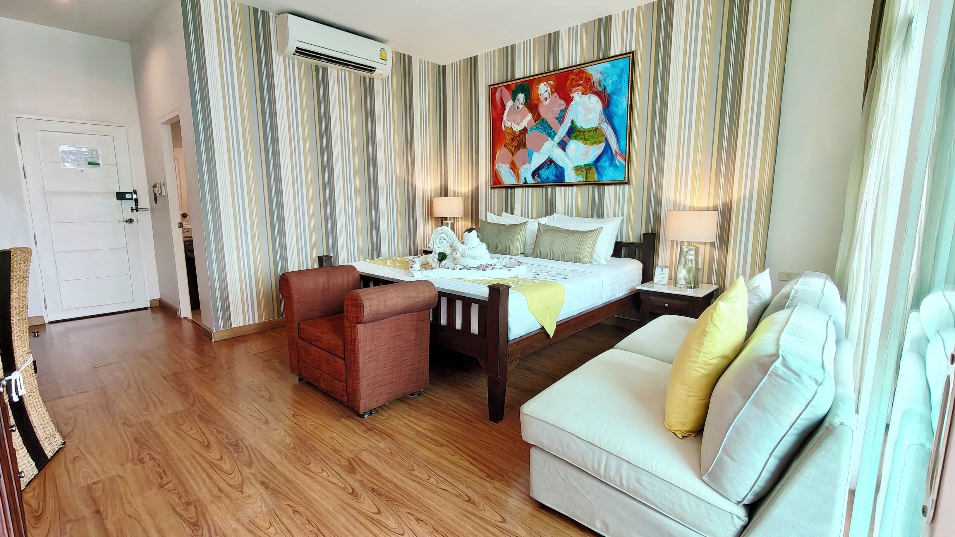 A hotel room with a bed, a sofa, a chair, a painting, and an air conditioning unit.