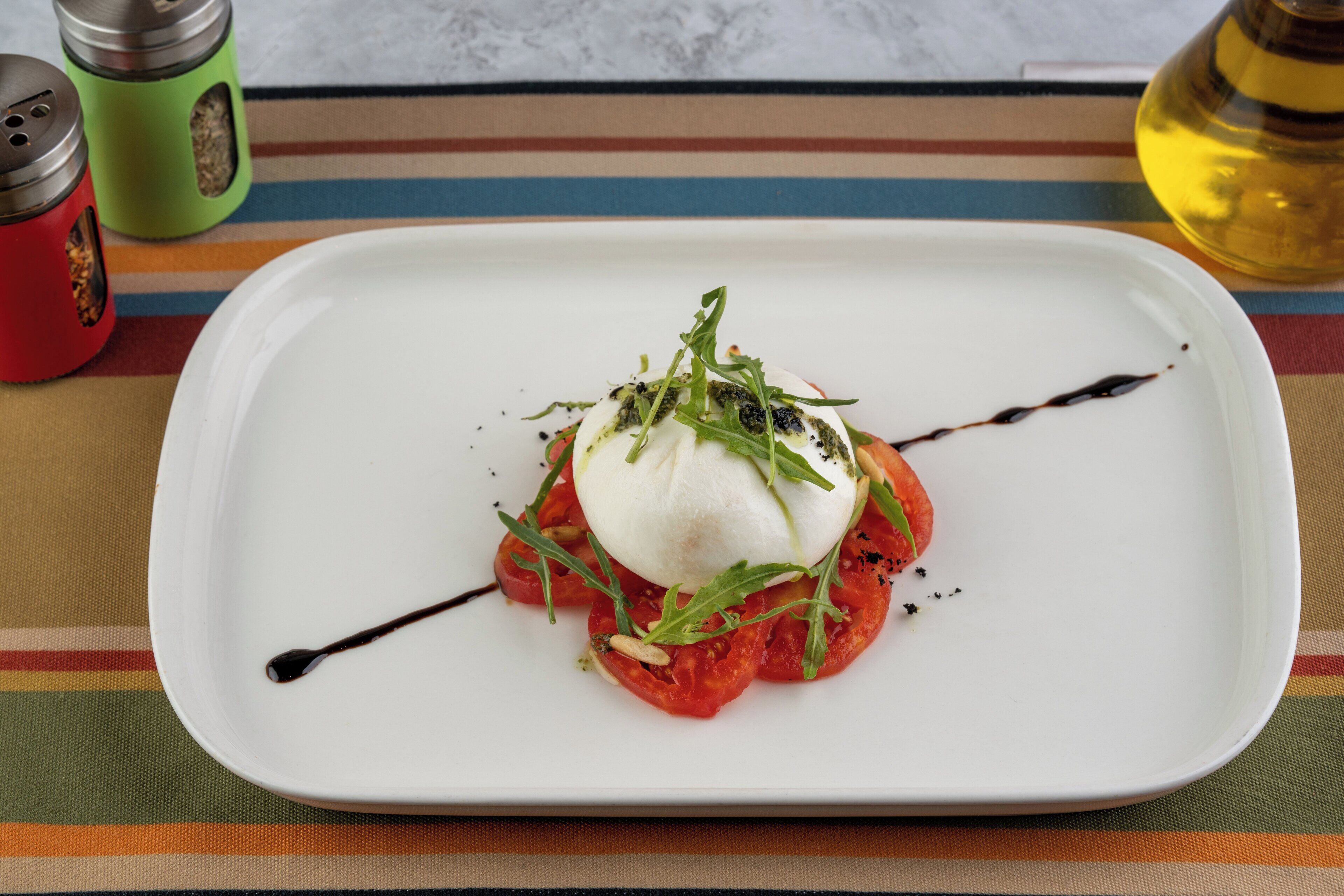 Ein weißer Teller mit Tomatensalat, belegt mit Mozzarella und Rucola, mit einem Soßenüberzug und einer schwarzen Linie als Dekoration.