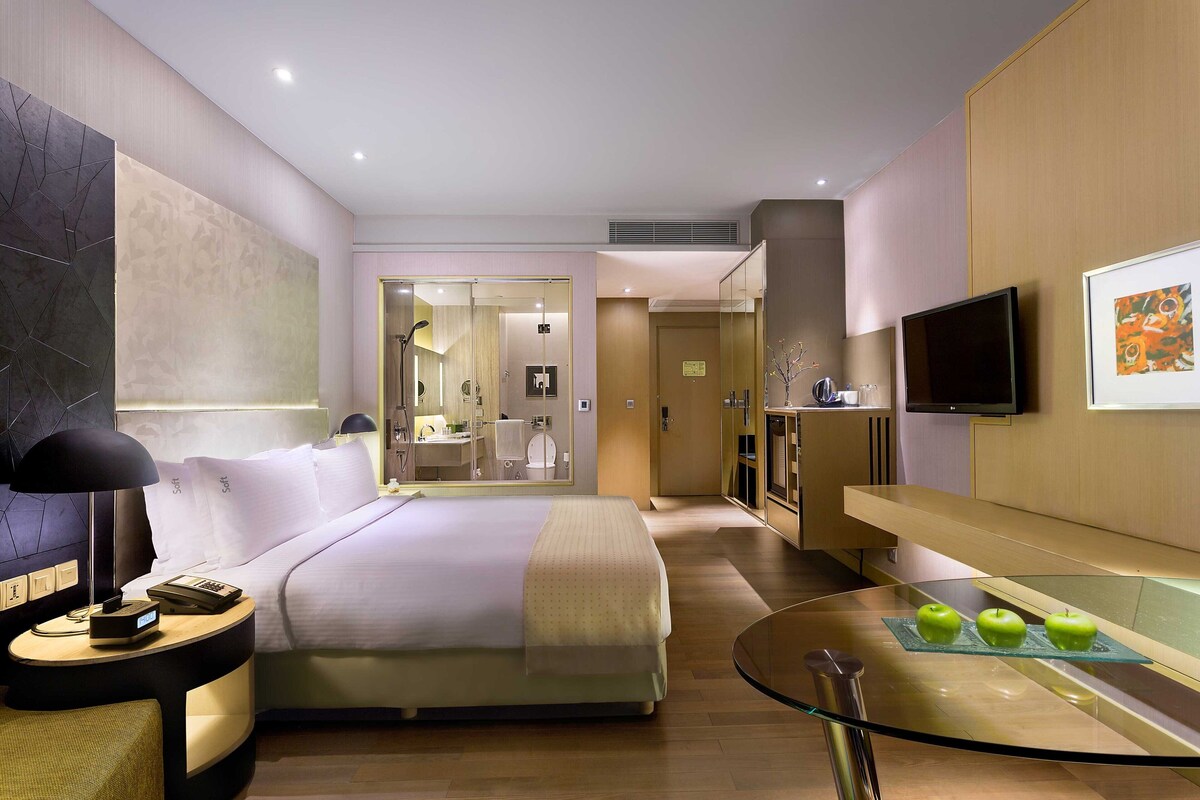 Een moderne hotelkamer met een groot bed, nachtkastje met een lamp, een glazen tafel met appels, een televisie, en een badkamer zichtbaar door een glazen deur.