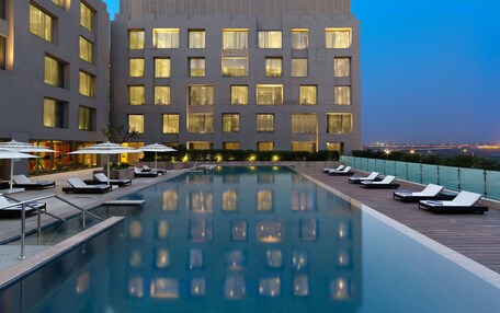 A hotel pool area with sun loungers and umbrellas, surrounded by a building with multiple windows and a view of the cityscape at dusk.