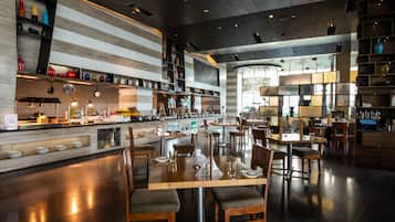 Un restaurante moderno con zona de bar, mesas y sillas de madera, y una variedad de utensilios de cocina y adornos en estantes.
