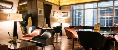 A modern hotel lobby with a wooden floor, leather chairs, a glass table, and a large window providing a view of the city.