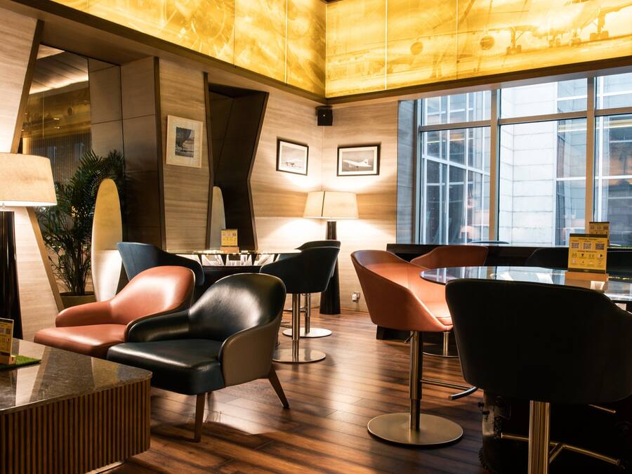 Um saguão de hotel moderno com piso de madeira, poltronas de couro, uma mesa de vidro e uma janela ampla com vista para a cidade.