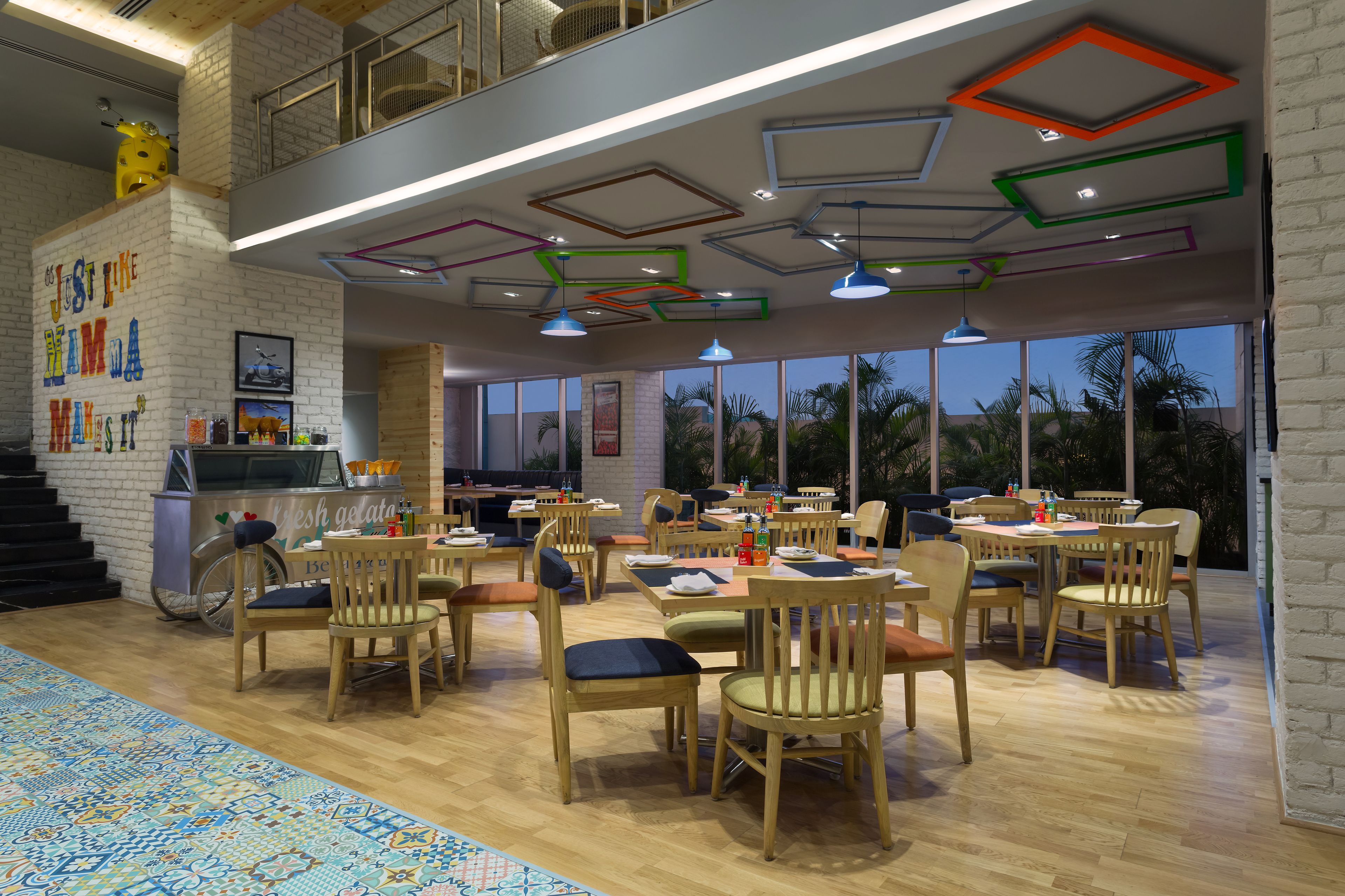 A spacious dining area with wooden chairs and tables, a high ceiling with geometric light fixtures, and large windows offering a view of palm trees.