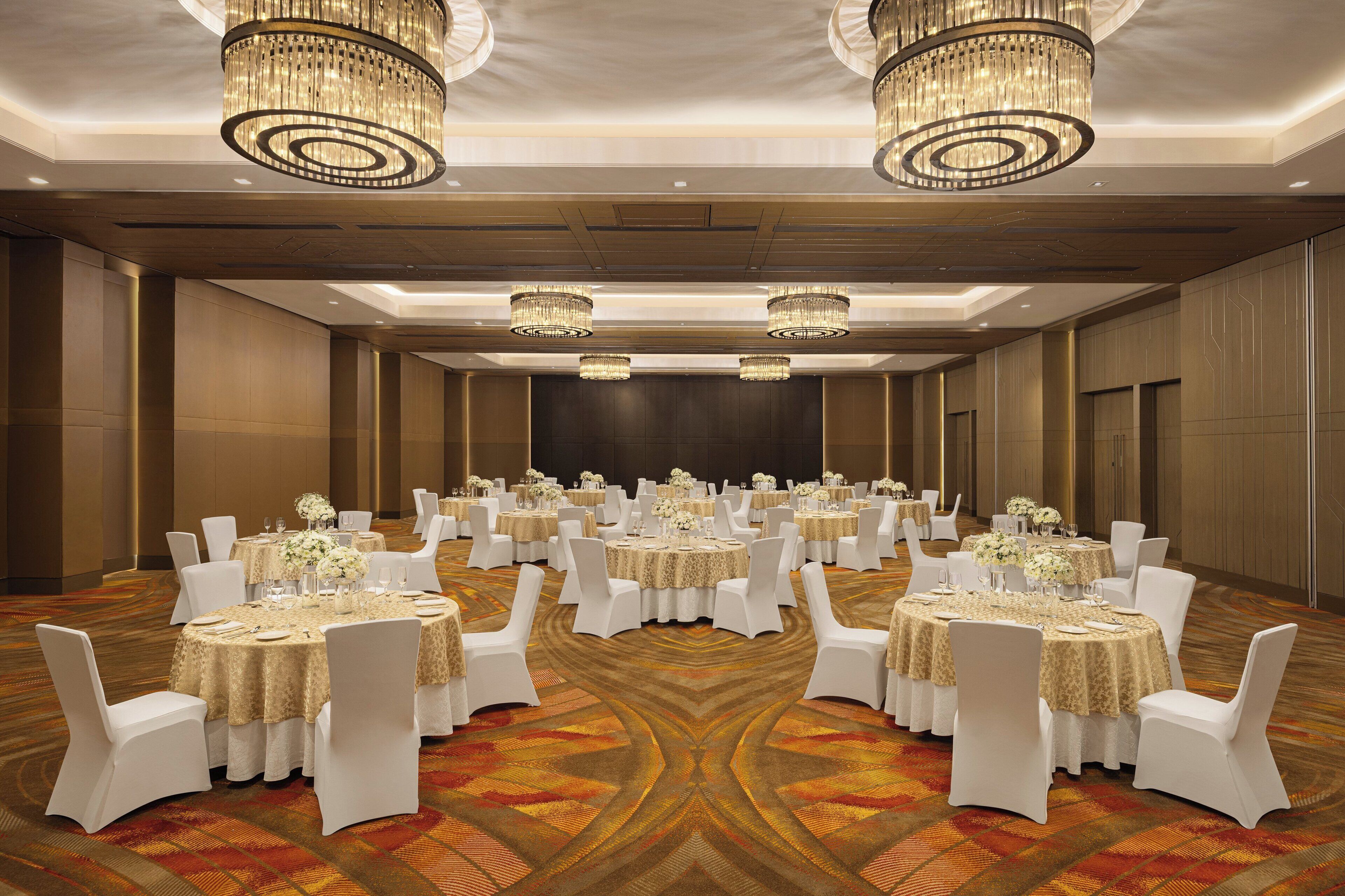 A banquet hall with round tables, white chairs, chandeliers, and a patterned carpet.