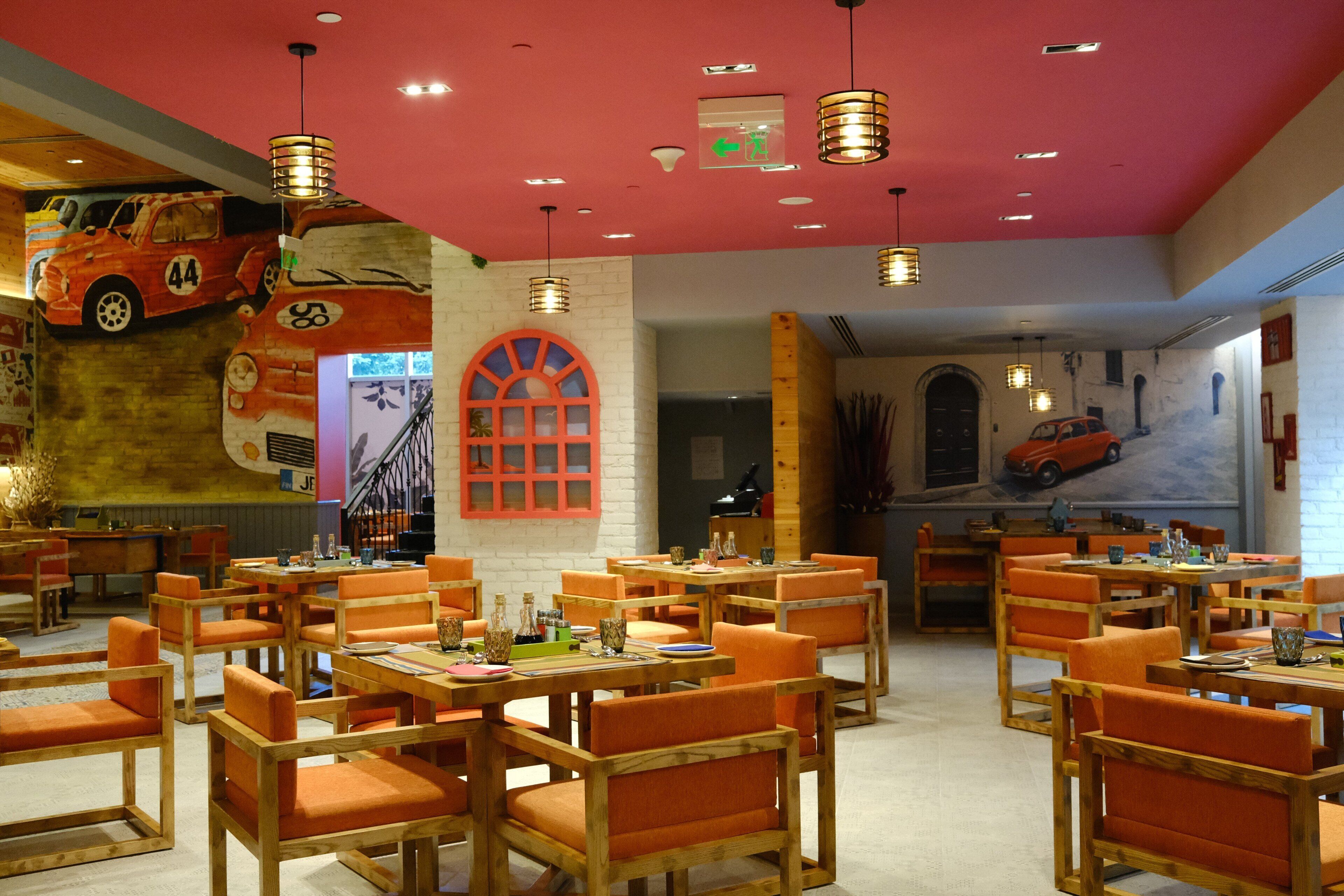 Un restaurante con mesas de madera y sillas anaranjadas, techo rojo y un mural de un coche en la pared.
