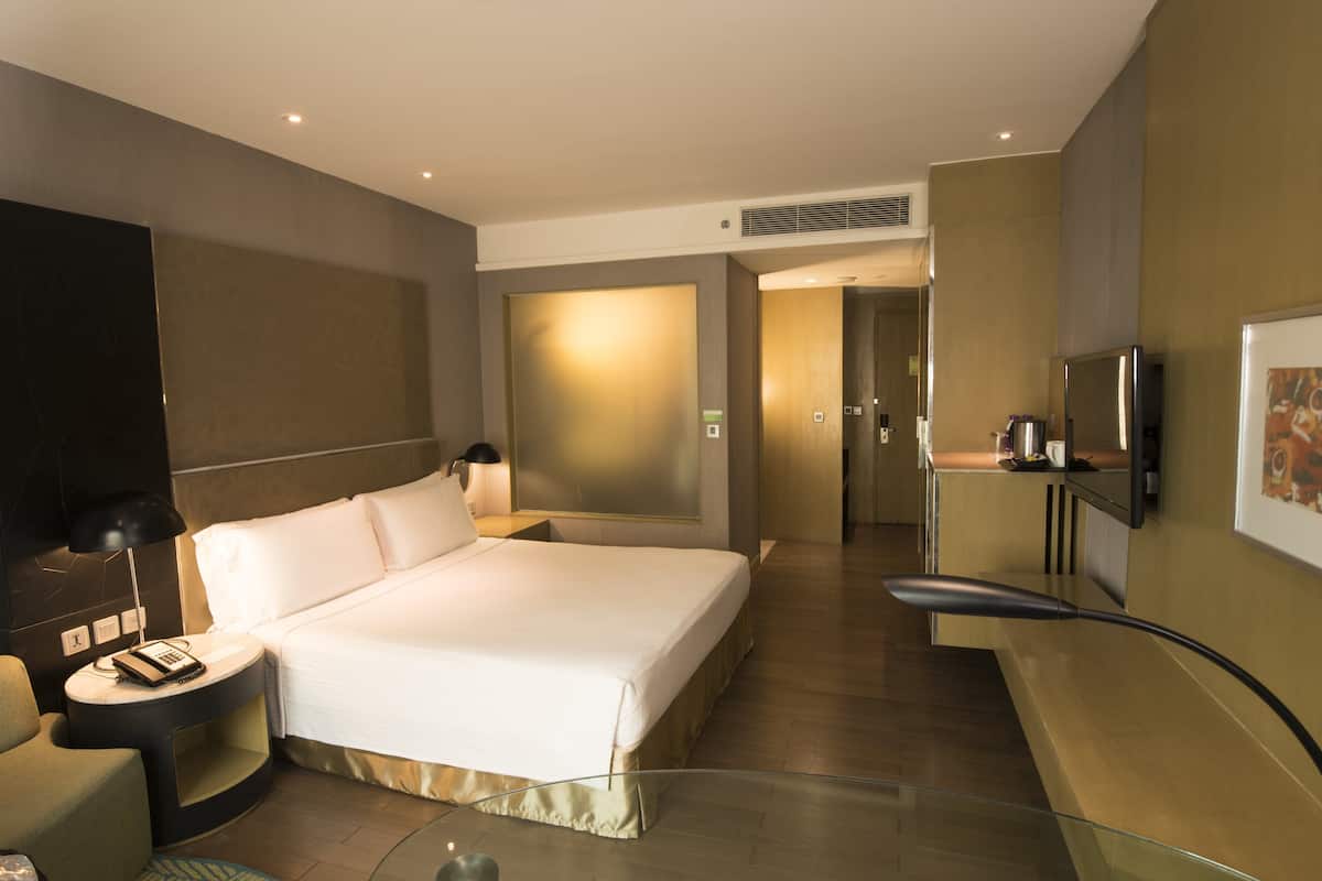 A modern hotel room with a large bed, bedside table with a lamp, a flat-screen TV mounted on the wall, and a small kitchenette area.
