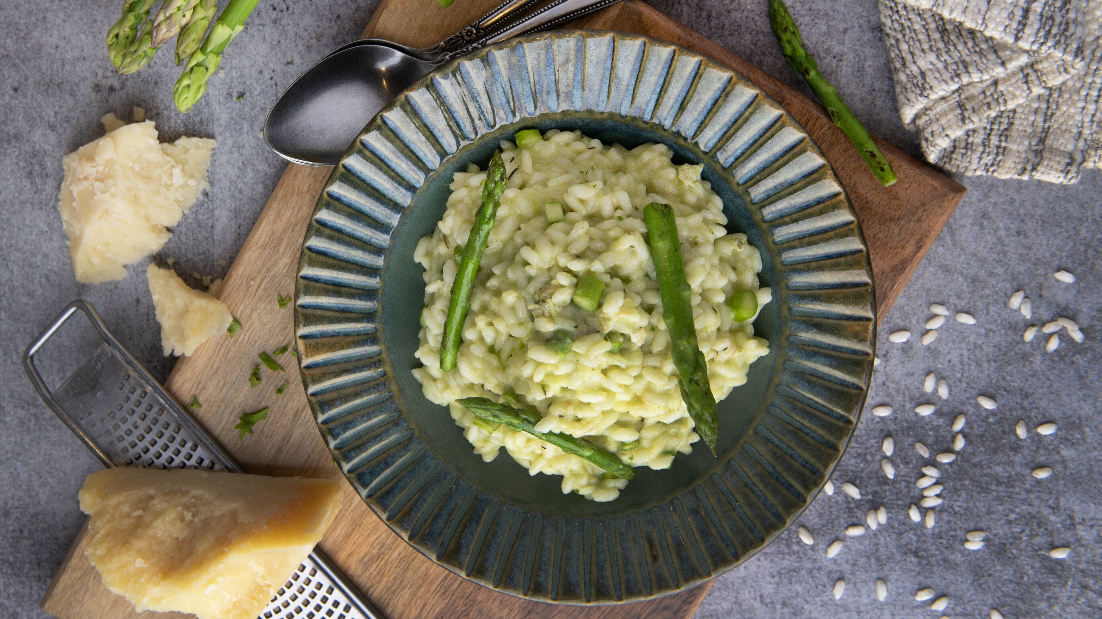 Un plato de risotto con espárragos sobre un plato de cerámica, acompañado de un rallador, una cuchara y un poco de pan en una tabla de madera.
