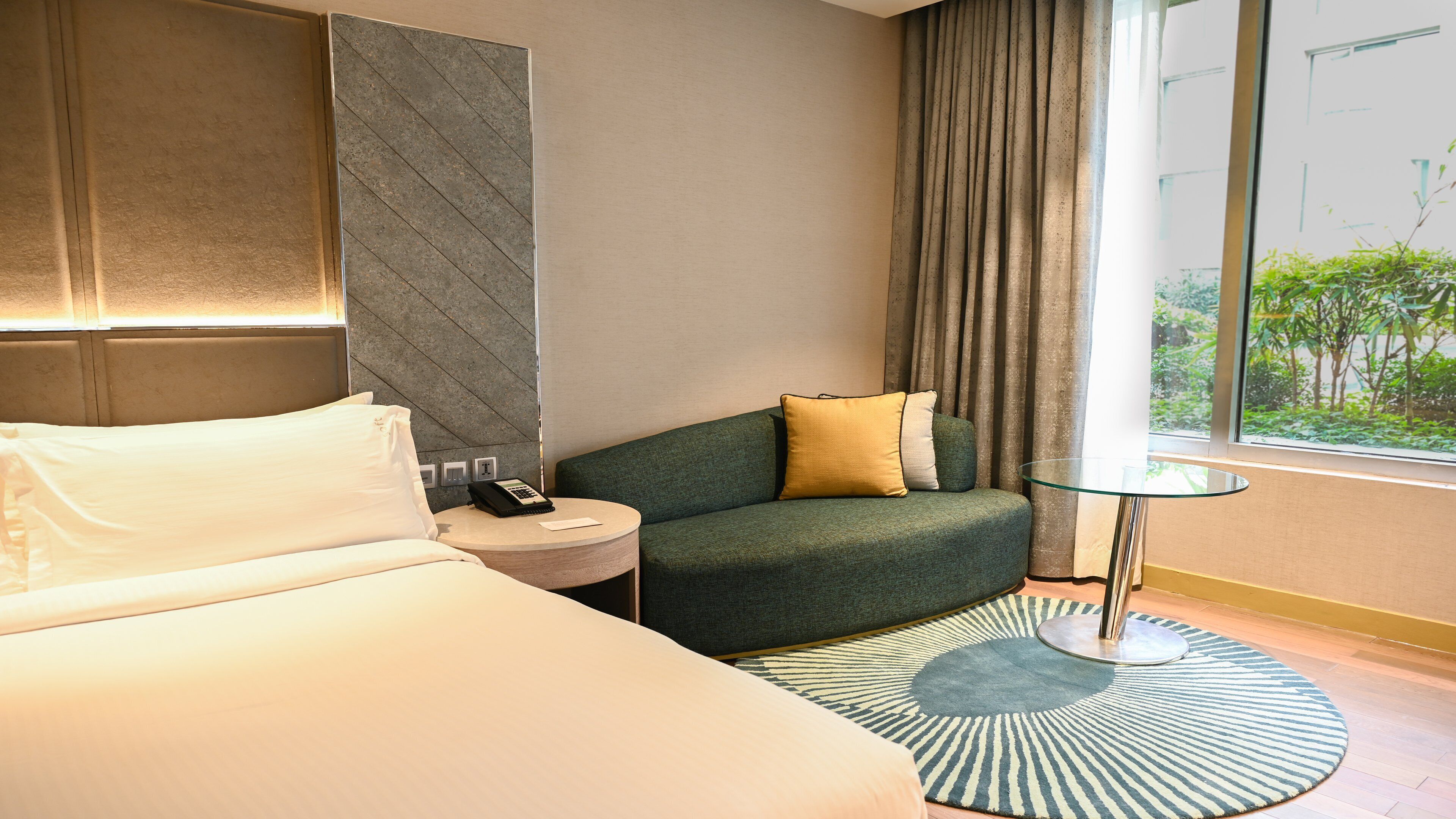 A hotel room with a bed, a small round table with a phone, a green sofa with a yellow pillow, a patterned rug, and a window with a view of greenery.