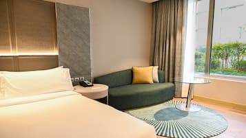 A hotel room with a bed, a small round table with a phone, a green sofa with a yellow pillow, a patterned rug, and a window with a view of greenery.