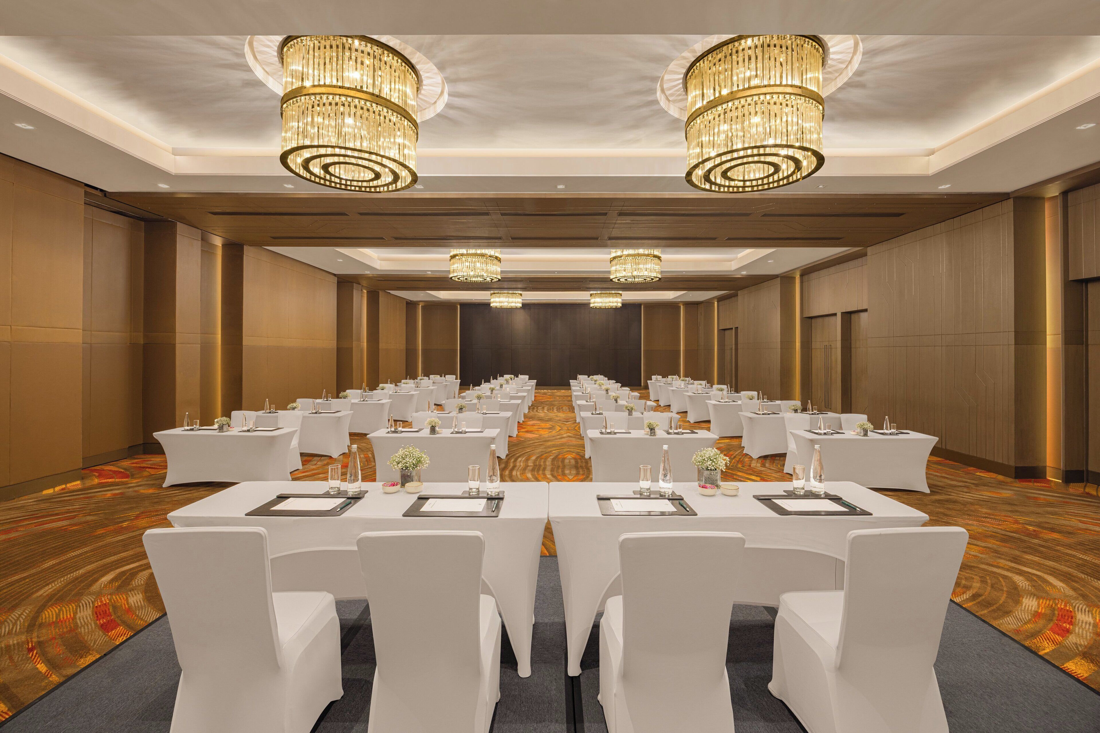 A conference room with round tables, white chairs, chandeliers, and a carpeted floor.