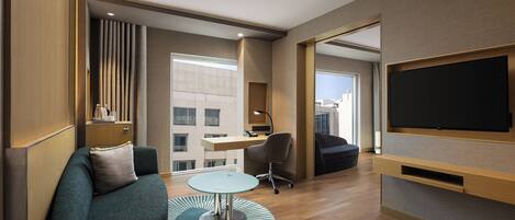 A modern hotel room with a sofa, a small round table, a desk, a chair, and a TV mounted on the wall.