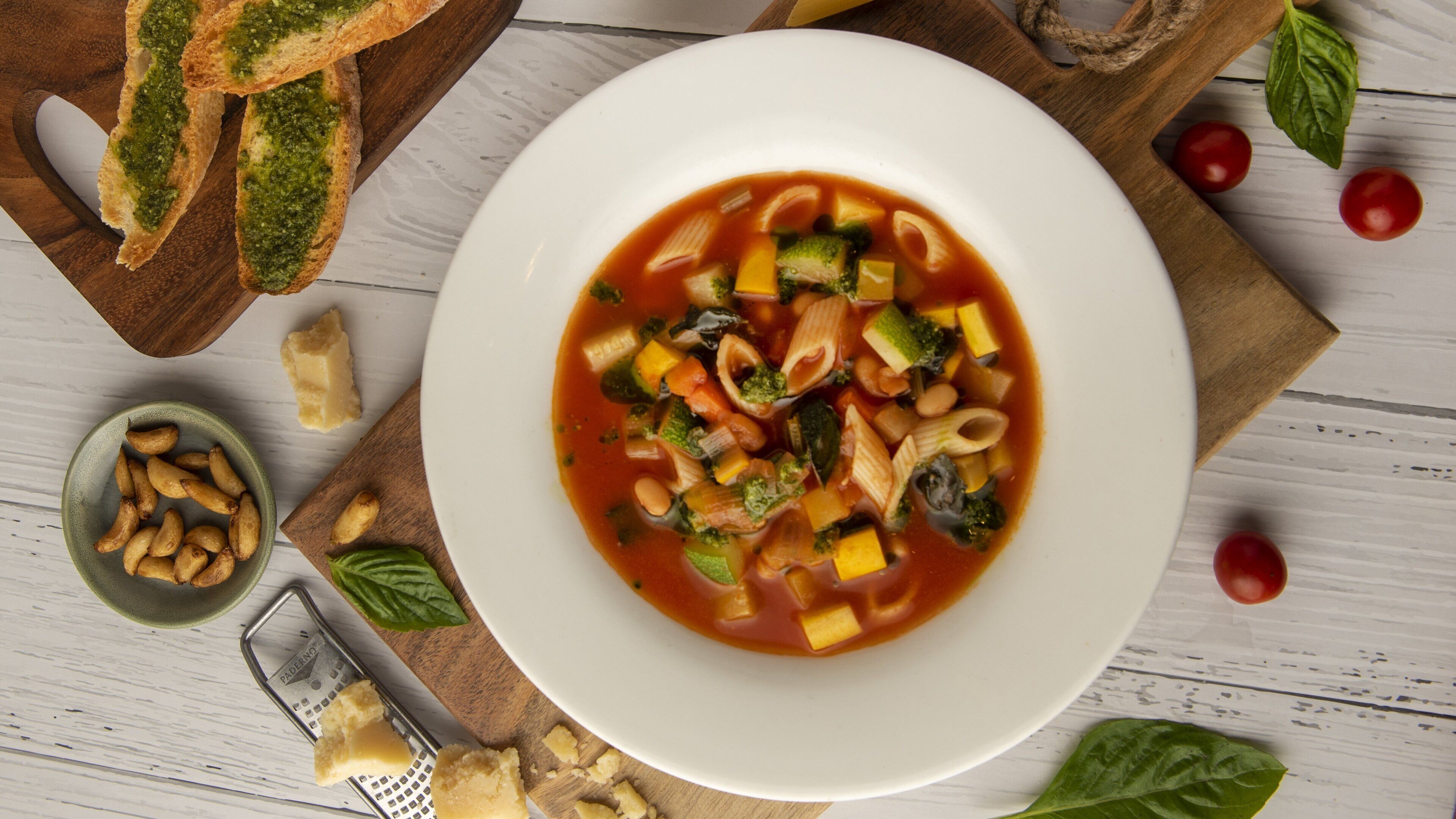 A bowl of vegetable soup with pasta, surrounded by bread, tomatoes, and basil leaves on a wooden table.