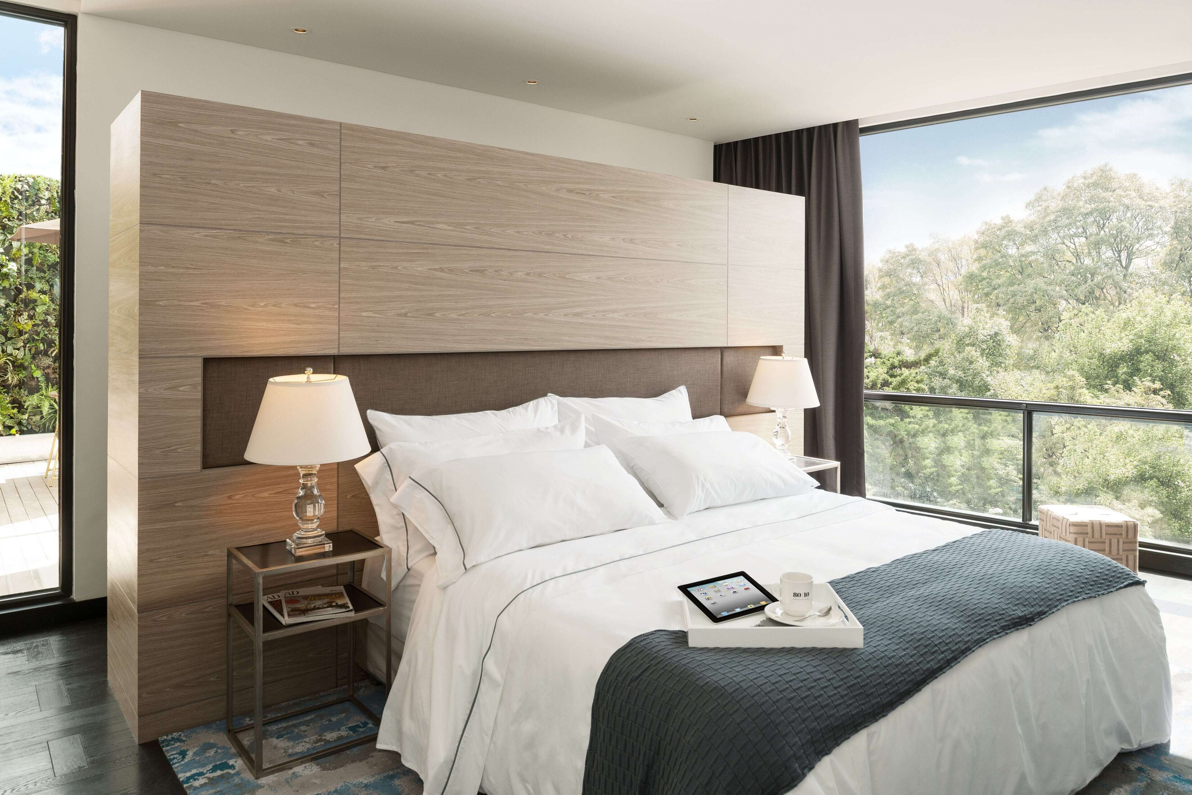 A modern hotel room with a large bed, a bedside table with a lamp, a bedside table with a tray, and a view of trees through large windows.