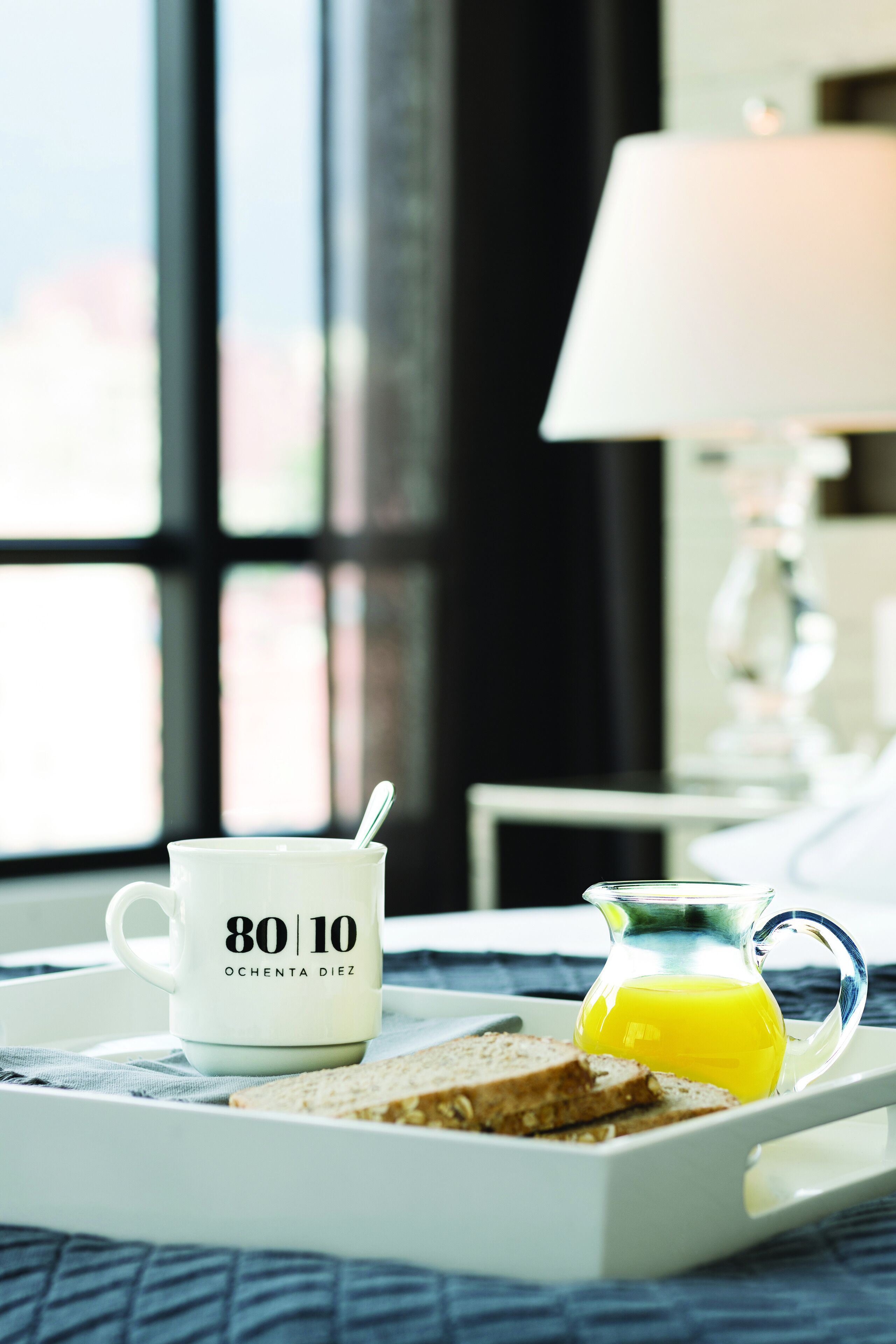 A breakfast tray with a mug labeled "80 | 10" and a glass pitcher of orange juice on a bed with a view of a cityscape through large windows.